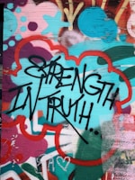 Graffiti art featuring bold black lettering of the words 'Strength in Truth' against a vibrant, multicolored background. The background includes abstract shapes and patterns in shades of teal, red, pink, and orange, creating a dynamic and energetic visual effect. The surface appears to be a wooden panel with visible grain.