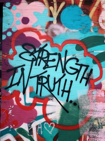 Graffiti art featuring bold black lettering of the words 'Strength in Truth' against a vibrant, multicolored background. The background includes abstract shapes and patterns in shades of teal, red, pink, and orange, creating a dynamic and energetic visual effect. The surface appears to be a wooden panel with visible grain.
