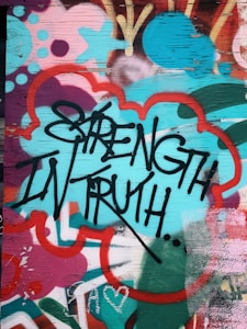 Graffiti art featuring bold black lettering of the words 'Strength in Truth' against a vibrant, multicolored background. The background includes abstract shapes and patterns in shades of teal, red, pink, and orange, creating a dynamic and energetic visual effect. The surface appears to be a wooden panel with visible grain.