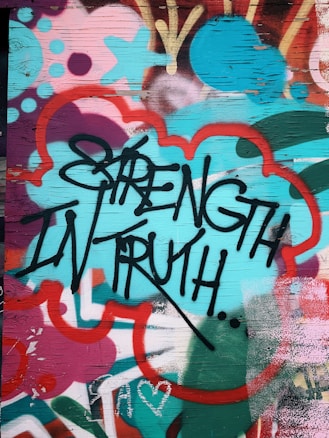 Graffiti art featuring bold black lettering of the words 'Strength in Truth' against a vibrant, multicolored background. The background includes abstract shapes and patterns in shades of teal, red, pink, and orange, creating a dynamic and energetic visual effect. The surface appears to be a wooden panel with visible grain.