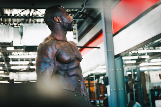 A muscular man with tattoos stands in a gym, highlighted by ceiling lights and a background of gym equipment and mirrors. He appears to be focused and possibly preparing for a workout.