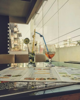 Two glasses sit on a table, one filled with a mojito garnished with a lime slice and the other with a partially empty orange drink. A menu is spread open in front of them. The setting appears to be a modern cafe or restaurant with a glass balcony offering a view of urban buildings in the background.