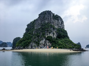 A large, lush, rocky island rises from calm waters surrounded by smaller landforms in the distance. The island features dense greenery and steep, rugged rock faces. A small sandy beach is visible at the base, with colorful kayaks lined up on the shore.