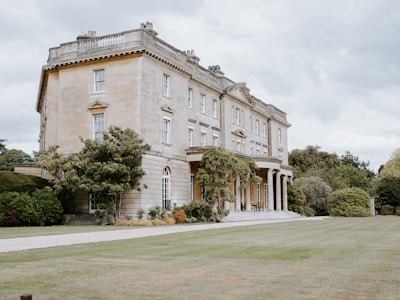 An elegant, large two-story stone mansion with classical architectural elements stands amidst manicured gardens and lush greenery. The facade features large windows and a formal entrance with columns. The surrounding greenery includes trees, shrubs, and a well-maintained lawn.