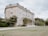 An elegant, large two-story stone mansion with classical architectural elements stands amidst manicured gardens and lush greenery. The facade features large windows and a formal entrance with columns. The surrounding greenery includes trees, shrubs, and a well-maintained lawn.