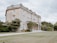 An elegant, large two-story stone mansion with classical architectural elements stands amidst manicured gardens and lush greenery. The facade features large windows and a formal entrance with columns. The surrounding greenery includes trees, shrubs, and a well-maintained lawn.
