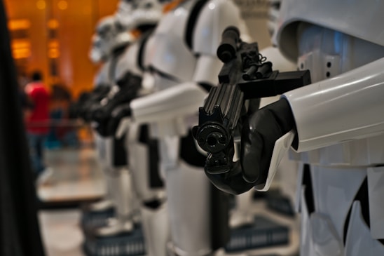 A row of futuristic soldiers in white armor with black gloves, holding black weapons. The focus is on the foreground soldier's weapon, highlighting details and edges sharply, while the background soldiers appear slightly blurred.
