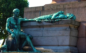 A bronze sculpture depicts a seated male figure with draped clothing, holding the arm of a reclining female figure on a stone platform. The green patina of the bronze stands out against the stone backdrop and the natural setting.