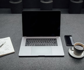 turned-off MacBook Pro between cup of coffee, iPhone, notebook, and pen