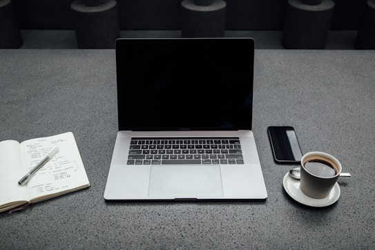 A neatly arranged workspace on a gray textured surface featuring a laptop with a blank screen, an open notebook with a pen placed on it, a smartphone, and a cup of coffee on a saucer with a spoon.