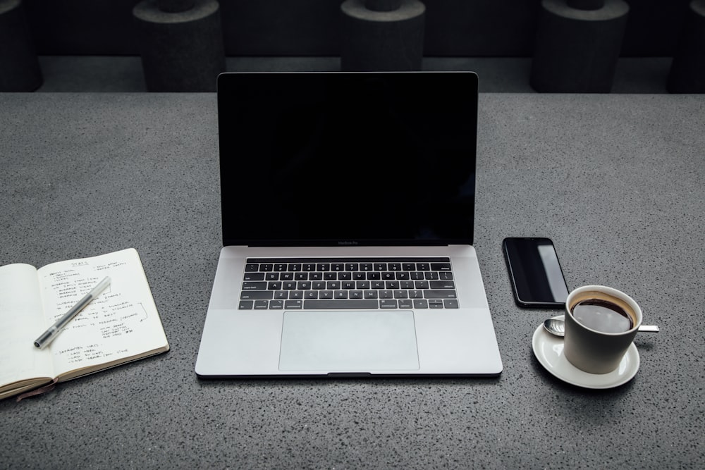 turned-off MacBook Pro between cup of coffee, iPhone, notebook, and pen