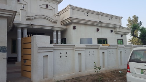 A white, single-story house with an elaborate entrance featuring columns and decorative architectural elements. Surrounding the house is a high boundary wall with geometric patterns. A white vehicle is parked in front of the house on a patch of dry grass, next to some small plants and a tree.