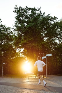 A jogger wearing a white shirt and black shorts is running along a paved path in a park. The sun is setting in the background, casting a warm, golden glow through the dense green trees. The path is bordered by wooden posts, and there's a street sign nearby.
