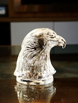 A crystal sculpture of an eagle's head is displayed on a reflective surface. The intricate details of the glass capture the feathers' texture, beautifully refracting light to create a shimmering effect.