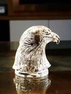 A crystal sculpture of an eagle's head is displayed on a reflective surface. The intricate details of the glass capture the feathers' texture, beautifully refracting light to create a shimmering effect.