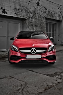 A red car with a prominent front grille and the Mercedes-Benz emblem parked in front of a weathered, industrial building. The background features concrete walls with patches of peeling paint and two large metal doors.