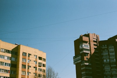 Two mid-century residential apartment buildings under a clear blue sky. The left building is a light yellow with a flat roof, while the right building is a dark red brick with multiple protrusions. Leafless trees are visible at the bottom.