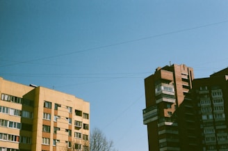 Two mid-century residential apartment buildings under a clear blue sky. The left building is a light yellow with a flat roof, while the right building is a dark red brick with multiple protrusions. Leafless trees are visible at the bottom.