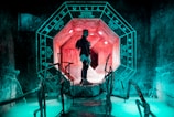 A person stands inside an illuminated, futuristic chamber with a prominent octagonal shape. The walls are adorned with glowing symbols resembling ancient or mystical characters, while the chamber emits an eerie teal glow. A pathway with handrails leads towards the center, surrounded by luminescent blue water.