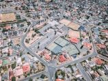 Aerial view of a suburban area featuring a network of roads, residential houses, and a sports complex. The sports complex includes multiple tennis courts, parking lots, and surrounding greenery. Houses are neatly arranged along the streets, displaying a variety of colors including red, gray, and white roofs.