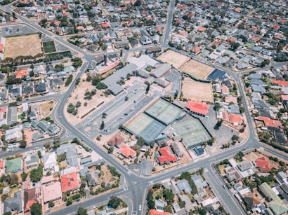Aerial view of a suburban area featuring a network of roads, residential houses, and a sports complex. The sports complex includes multiple tennis courts, parking lots, and surrounding greenery. Houses are neatly arranged along the streets, displaying a variety of colors including red, gray, and white roofs.