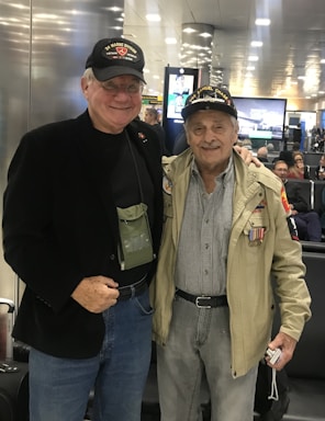 Two elderly men are standing side by side at an airport terminal. Both are wearing dark baseball caps with military insignias. The man on the left is dressed in a black jacket and jeans, while the man on the right wears a light-colored jacket adorned with medals and a checkered shirt. They are smiling, and the ambience suggests a warm interaction. Behind them, other travelers are seated, and there are digital displays and bright overhead lights in the background.
