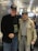 Two elderly men are standing side by side at an airport terminal. Both are wearing dark baseball caps with military insignias. The man on the left is dressed in a black jacket and jeans, while the man on the right wears a light-colored jacket adorned with medals and a checkered shirt. They are smiling, and the ambience suggests a warm interaction. Behind them, other travelers are seated, and there are digital displays and bright overhead lights in the background.