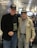 Two elderly men are standing side by side at an airport terminal. Both are wearing dark baseball caps with military insignias. The man on the left is dressed in a black jacket and jeans, while the man on the right wears a light-colored jacket adorned with medals and a checkered shirt. They are smiling, and the ambience suggests a warm interaction. Behind them, other travelers are seated, and there are digital displays and bright overhead lights in the background.