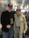 Two elderly men are standing side by side at an airport terminal. Both are wearing dark baseball caps with military insignias. The man on the left is dressed in a black jacket and jeans, while the man on the right wears a light-colored jacket adorned with medals and a checkered shirt. They are smiling, and the ambience suggests a warm interaction. Behind them, other travelers are seated, and there are digital displays and bright overhead lights in the background.