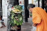 An elderly person dressed in an orange garment is leaning toward a column adorned with an elaborate arrangement of green leaves and colorful flowers, including yellow and white ones. The surrounding area appears to be a temple with ornate carvings and pillars.