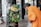 An elderly person dressed in an orange garment is leaning toward a column adorned with an elaborate arrangement of green leaves and colorful flowers, including yellow and white ones. The surrounding area appears to be a temple with ornate carvings and pillars.