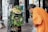 An elderly person dressed in an orange garment is leaning toward a column adorned with an elaborate arrangement of green leaves and colorful flowers, including yellow and white ones. The surrounding area appears to be a temple with ornate carvings and pillars.