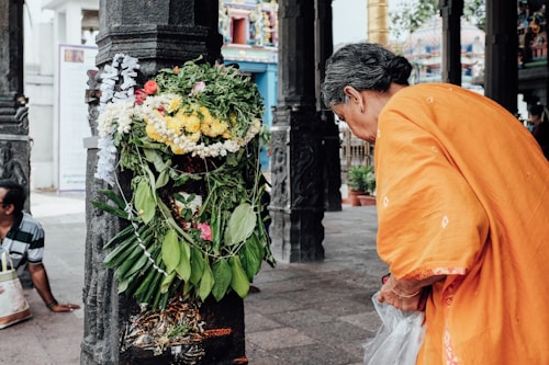 An elderly person dressed in an orange garment is leaning toward a column adorned with an elaborate arrangement of green leaves and colorful flowers, including yellow and white ones. The surrounding area appears to be a temple with ornate carvings and pillars.