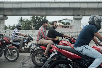 Several people ride motorcycles on a road alongside a concrete bridge. Two men are on a red bike, with one wearing a plaid shirt and the other in a black shirt. Another man in a helmet rides a motorcycle nearby. The environment includes concrete structures, a brick wall, and some trees in the background.
