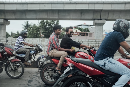 Several people ride motorcycles on a road alongside a concrete bridge. Two men are on a red bike, with one wearing a plaid shirt and the other in a black shirt. Another man in a helmet rides a motorcycle nearby. The environment includes concrete structures, a brick wall, and some trees in the background.