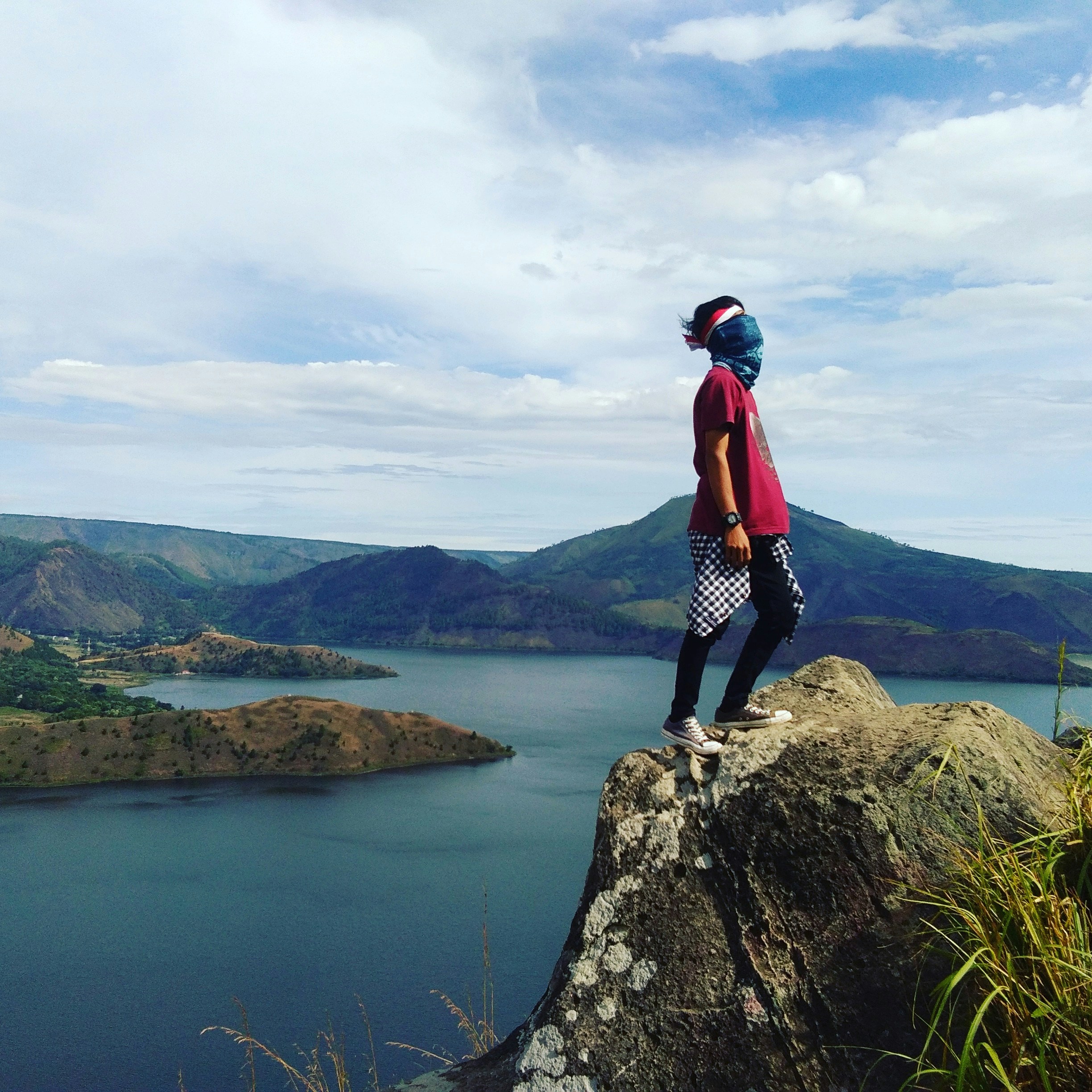 Orang yang mengenakan kemeja merah berdiri di atas batu abu-abu di seberang gunung