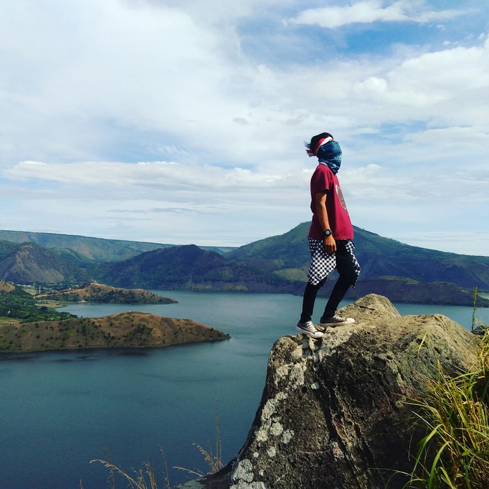 person wearing red shirt standing on gray rock across mountain