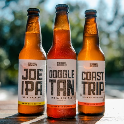 Three beer bottles stand on a wooden surface with condensation indicating they are chilled. The labels on the bottles read Joe IPA, Goggle Tan, and Coast Trip, each offering different beer types. Sunlight illuminates the bottles, creating a refreshing and inviting appearance.