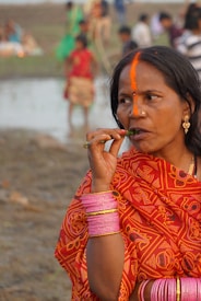 A woman is dressed in vibrant traditional attire, wearing ornate jewelry and pink bangles. She appears to be outdoors, with a blurred background showing people near a body of water. An orange mark adorns her forehead.