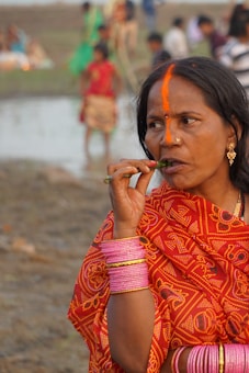 A woman is dressed in vibrant traditional attire, wearing ornate jewelry and pink bangles. She appears to be outdoors, with a blurred background showing people near a body of water. An orange mark adorns her forehead.