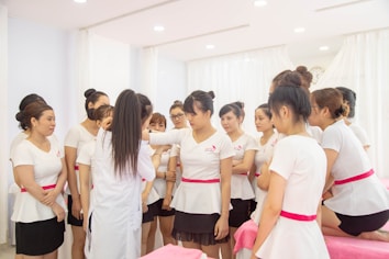 A group of women wearing uniforms with white tops and pink sashes is gathered indoors. They appear to be in a training or instructional session, with one woman demonstrating something to the others. The room is bright with soft lighting and white curtains, conveying a clean and professional environment.