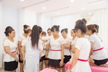 A group of women wearing uniforms with white tops and pink sashes is gathered indoors. They appear to be in a training or instructional session, with one woman demonstrating something to the others. The room is bright with soft lighting and white curtains, conveying a clean and professional environment.