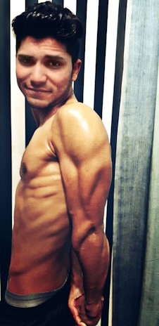 A muscular man is standing sideways, showcasing his well-defined arms and upper body. He is situated in front of a background featuring alternating black and white stripes and a gray curtain on the right.