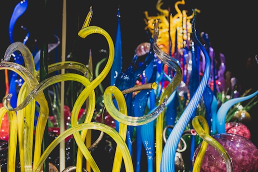 Vibrant glass sculptures rise in twisting, organic forms with vivid colors blending seamlessly. The intricate design showcases a range of translucent tubes and spheres, reflecting light to create a dynamic visual experience.