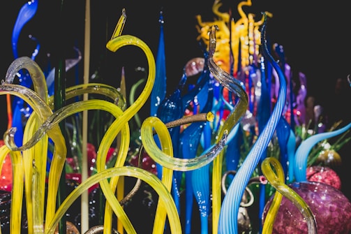 Vibrant glass sculptures rise in twisting, organic forms with vivid colors blending seamlessly. The intricate design showcases a range of translucent tubes and spheres, reflecting light to create a dynamic visual experience.