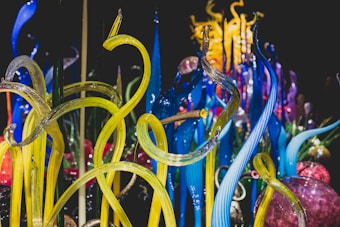 Vibrant glass sculptures rise in twisting, organic forms with vivid colors blending seamlessly. The intricate design showcases a range of translucent tubes and spheres, reflecting light to create a dynamic visual experience.