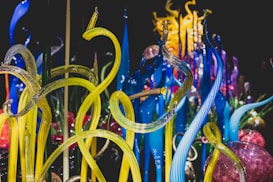 Vibrant glass sculptures rise in twisting, organic forms with vivid colors blending seamlessly. The intricate design showcases a range of translucent tubes and spheres, reflecting light to create a dynamic visual experience.