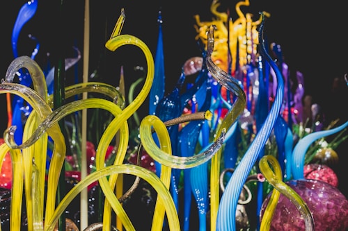 Vibrant glass sculptures rise in twisting, organic forms with vivid colors blending seamlessly. The intricate design showcases a range of translucent tubes and spheres, reflecting light to create a dynamic visual experience.
