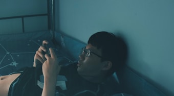 A person wearing glasses is lying on a bed, holding a smartphone. The room is dimly lit, creating a calm and relaxed atmosphere. The bed has a star-patterned bedsheet, and the overall color tone is blue-green.