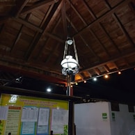 A traditional wooden ceiling with a hanging decorative lamp is featured, accompanied by a bulletin board displaying announcements and information in a public space. Colored lights are strung along near the ceiling.